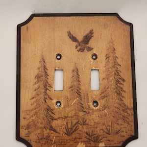 Peut inclure: Une plaque d'interrupteur double en bois rectangulaire avec une bordure brun foncé et un motif pyrogravé représentant un aigle en vol au-dessus d'un paysage forestier avec trois pins et de la végétation basse, incluant quatre trous de vis.
