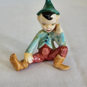 Könnte beinhalten: Eine Keramikfigur eines sitzenden Elfen. Der Elf trägt einen grünen Hut, eine blau-grüne Jacke mit schwarzen Knöpfen und eine rote Hose. Die Stiefel des Elfen sind beige, und er hat einen nachdenklichen Ausdruck.