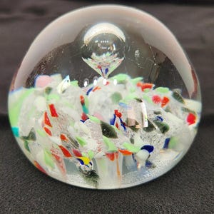 May include: A clear glass paperweight with colourful, abstract inclusions. The paperweight has a rounded shape with a bubble-like feature at the top. The inclusions are in shades of red, green, blue, and white, creating a decorative effect.