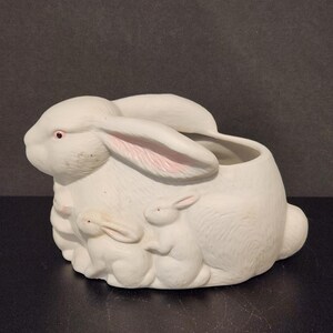 May include: A white ceramic planter shaped like a large rabbit with two smaller rabbits sculpted on the side. The large rabbit features pink-tinted inner ears and pink eyes, with a hollow opening at the top for plants or decorative items.