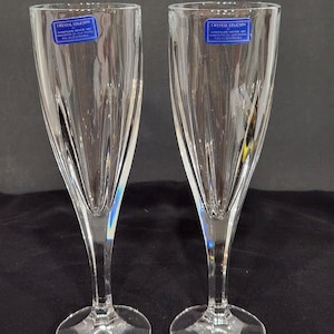 May include: Two clear crystal champagne flutes with a vertical, ridged design. Each glass has a long stem and a round base. Blue and white labels with the text "CRYSTAL LEGENDS" are affixed to the top of each glass.