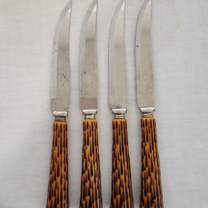 May include: Four vintage steak knives with stainless steel blades and handles with a brown and black tortoiseshell pattern. The knives are arranged vertically on a white surface.