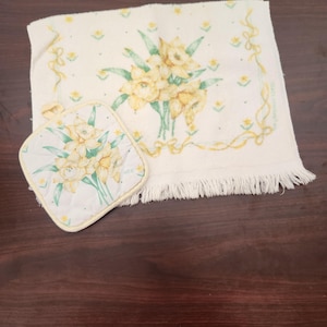 May include: A cream-colored rectangular hand towel with fringe and a matching square quilted potholder. Both items feature a printed design of yellow daffodils with green leaves and a yellow ribbon border pattern on a white background.