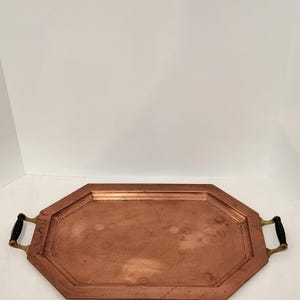 May include: Copper-colored octagonal tray with black and brass handles. The tray has a slightly aged appearance, with visible patina. The handles are black with brass accents. A decorative serving piece.