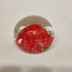 May include: A clear glass paperweight with a vibrant red, textured interior. The paperweight is spherical, with small air bubbles visible within the glass. The red interior has a layered appearance, creating depth and visual interest.