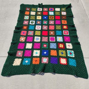 May include: A rectangular crochet blanket featuring a dark forest green border and a grid of multicolored granny squares. The squares are arranged in rows and columns, displaying a variety of vibrant hues including red, blue, pink, yellow, and white.