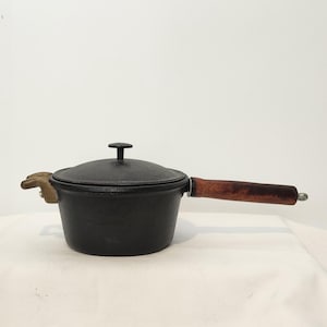 May include: A black cast iron saucepan with a matching lid featuring a central knob. The pot has a long, cylindrical wooden handle with a dark brown finish and a small, light-colored metal helper handle on the opposite side of the main handle.