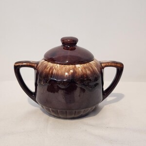May include: A brown ceramic sugar bowl with two handles and a matching lid. The piece features a dark brown base with a lighter, cream-colored drip glaze pattern around the upper rim and lid, and vertical ribbed detailing along the bottom section.