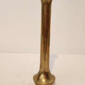 May include: A tall, slender brass bud vase with a polished metallic gold finish. The vase features a narrow, cylindrical neck that flares slightly at the top rim and expands into a rounded, bulbous base with a flat, circular bottom edge.