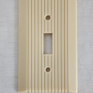Puede incluir: Cubierta de interruptor de luz rectangular de color crema con detalles verticales acanalados. La cubierta tiene una abertura rectangular en el centro y dos aberturas circulares.