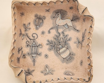 May include: A square ceramic dish with a textured, speckled beige surface and a scalloped edge. The interior features blue line drawings of a horse, a lyre, a lantern, a vase with foliage, and various small star and floral motifs.