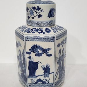 Puede incluir: Un tarro de cerámica blanco y azul con tapa. El tarro tiene forma octogonal y está decorado con ilustraciones azules de personas, dragones y motivos florales. La tapa también presenta diseños florales y de dragones azules.