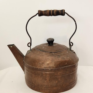 May include: A weathered copper kettle featuring a cylindrical body with horizontal ridges, a slanted spout, and a removable lid with a dark knob. A curved metal handle with a dark, textured wooden grip is attached to the top of the kettle.