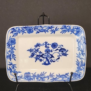 May include: A rectangular white ceramic tray featuring a blue floral pattern. The border is decorated with a continuous vine of flowers and leaves, while the center displays a symmetrical floral arrangement with stylized blossoms and foliage.