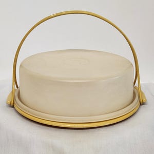 Könnte beinhalten: Ein cremefarbener Kuchenbehälter mit gelbem Boden und Griff. Der runde Behälter hat einen gewölbten Deckel und einen passenden gelben Griff, der sich über die Oberseite wölbt. Der Boden hat eine kontrastierende gelbe Farbe.