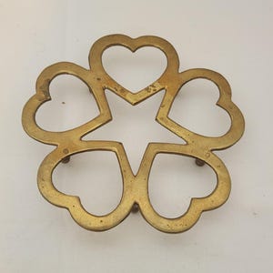 May include: A brass trivet featuring a symmetrical design of five interconnected heart shapes arranged in a circular pattern around a central star-shaped opening. The metal surface shows a weathered, golden-toned finish with visible patina.
