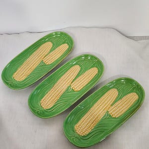 May include: Three ceramic serving platters with a bright green base and a detailed design of two ears of corn. The corn is yellow with textured kernels. The platters are oval-shaped.