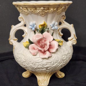 Może przedstawiać: Biały ceramiczny wazon z różową różą i niebieskimi kwiatami. Wazon ma ozdobny wzór i trzy nóżki.