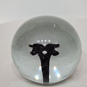 May include: A clear glass paperweight with a black abstract design inside. The paperweight is a sphere shape and has a slight grey tint. The design appears to be two figures connected by a stem.