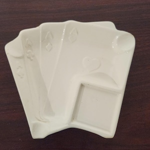Könnte beinhalten: Cremefarbene Keramikschale in Spielkartenform. Die Schale zeigt geprägte Kartensymbole: Karo, Kreuz und Herz. Die Schale ist für kleine Gegenstände konzipiert und vor einem dunkelbraunen Hintergrund platziert.