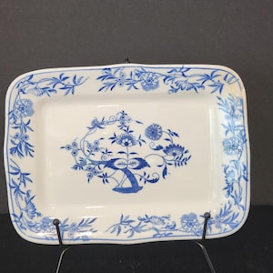 May include: A rectangular white ceramic serving tray featuring a blue floral pattern. The centre displays a detailed botanical motif, while the border is decorated with a continuous vine and flower design in matching blue tones.