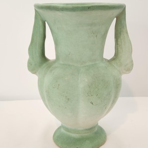 May include: A pale green ceramic vase featuring a wide, flared rim, a bulbous body with vertical fluted panels, and two symmetrical side handles. The surface displays a visible crackle glaze finish with subtle variations in colour intensity.