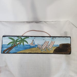 Könnte beinhalten: Eine rechteckige, handgemalte Strandszene auf einer Schiefertafel. Das Kunstwerk zeigt eine Palme, ein Segelboot, Liegestühle und das Meer. Die Tafel hat eine braune Lederschnur zum Aufhängen.