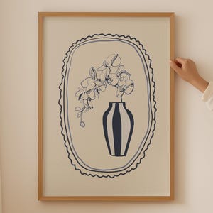 Puede incluir: Impresión artística enmarcada. La obra representa un jarrón a rayas azules y blancas con flores. La imagen está dentro de un óvalo festoneado azul sobre un fondo crema, enmarcado en madera clara.