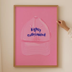 May include: Pink framed art print featuring a pink baseball cap illustration with the words "highly caffeinated" in blue. The artwork is displayed on a pink background. The frame is a light brown color.