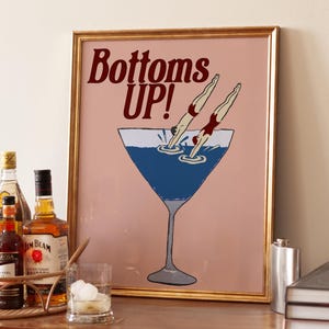 May include: A framed print with the words "Bottoms UP!" above a blue cocktail glass with two figures diving in. The print is gold-framed and sits on a wooden cabinet with bar accessories.