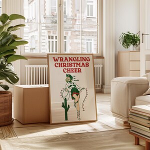 May include: A vintage-style poster with a woman in a green and white outfit holding a lasso with Christmas lights. The poster has the text "WRANGLING CHRISTMAS CHEER" in red and green letters. The woman is standing in front of a green cactus.
