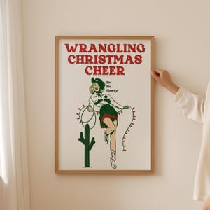May include: A framed print with a red and green illustration of a woman in a cowboy hat and skirt, holding a lasso with Christmas lights. The text "WRANGLING CHRISTMAS CHEER" is in red letters at the top of the print. The text "Ho Ho Howdy!" is in black letters below the woman.