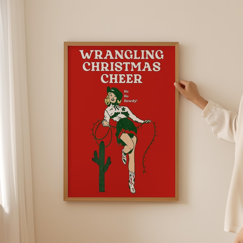 May include: A red poster with the text "WRANGLING CHRISTMAS CHEER" in white. A woman in a cowboy hat and outfit is holding a lasso with Christmas lights. The text "Ho Ho Howdy!" is below the woman.