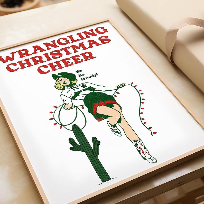 May include: A framed print with a green, red, and white illustration of a woman in a cowboy hat lassoing Christmas lights. The text "WRANGLING CHRISTMAS CHEER" is in red and green letters. The text "Ho Ho Howdy!" is in green letters.