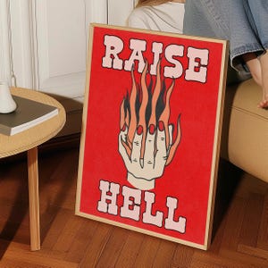 Könnte beinhalten: Ein gerahmter Kunstdruck mit rotem Hintergrund und den Worten "RAISE HELL" in rosa Buchstaben. Das Kunstwerk zeigt eine Hand mit langen roten Nägeln, mit Flammen, die von den Fingerspitzen aufsteigen. Der Rahmen ist hellholzfarben.