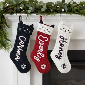 Op de afbeelding: Drie gebreide kerstsokken met kattengezichten en de namen: Carina, Everly en Honey. De sokken zijn zwart, rood en wit, met pootafdrukken aan de onderkant. Ze hangen aan een witte schoorsteenmantel versierd met groen.