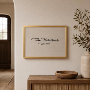 May include: A rectangular wall art piece featuring a gold frame and a light beige fabric background. The text "The Harrisons" is embroidered in black cursive script, with the text "Est. 2024" positioned directly underneath in a smaller font.