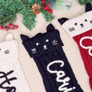 Op de afbeelding: Drie gebreide kerstsokken met kattengezichten. Eén is zwart met de naam "Cari" in wit, een andere is wit met een zwarte naam, en de derde is rood met een witte naam. De sokken hebben kabelbreipatronen en kattenoordetails.