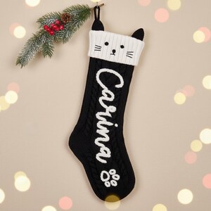 Op de afbeelding: Een zwart-witte gebreide kerstsok met een kattengezichtontwerp aan de bovenkant. De naam "Carina" is in witte letters op de voorkant geborduurd, met pootafdrukken aan de onderkant. Een takje dennen met rode bessen hangt erboven.