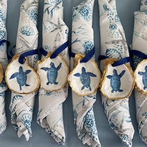 May include: Six white oyster shell napkin rings with gold trim and blue painted sea turtles. The napkin rings are tied with blue ribbon and are placed on white napkins with a blue and gray seashell pattern.