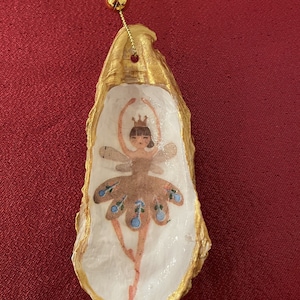 May include: A gold-painted oyster shell ornament with a painted fairy inside. The fairy is wearing a brown dress with blue accents and a crown.