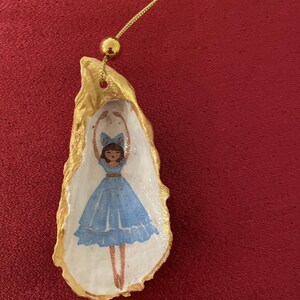 May include: A hand-painted oyster shell ornament featuring a ballerina in a blue dress and bow. The shell is edged in gold paint and suspended by a gold chain. The background is a deep red.