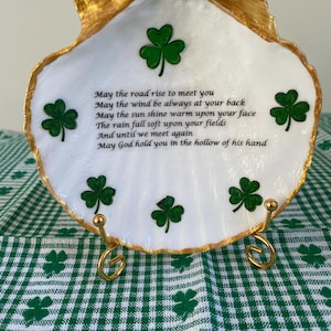 May include: White seashell with a gold rim and green shamrock designs. The seashell has an Irish blessing written on it: "May the road rise to meet you, May the wind be always at your back, May the sun shine warm upon your face, The rain fall soft upon your fields, And until we meet again, May God hold you in the hollow of his hand."