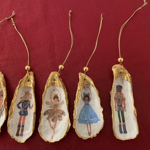 May include: Five gold-painted oyster shell ornaments with different Nutcracker-themed illustrations. The ornaments feature a ballerina, a nutcracker soldier, a mouse, and a fairy.
