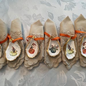 May include: Six white oyster shell napkin rings with gold trim and painted fall-themed gourds. Each ring is tied with an orange ribbon.