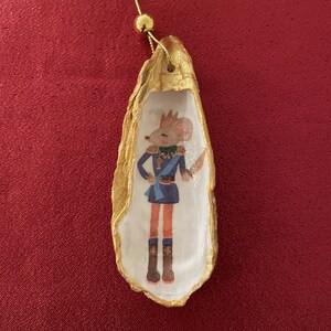 May include: A decorative Christmas ornament made from a gold-painted oyster shell. The shell contains a painted image of a mouse in a military uniform. The ornament hangs from a gold cord with a small gold bead.
