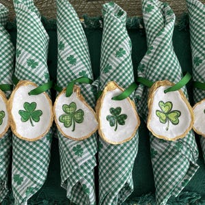 May include: Six green and white checkered napkins are arranged with oyster shell napkin rings. Each ring is white with a gold trim and a green shamrock design. Green ribbon is used to attach the rings to the napkins.