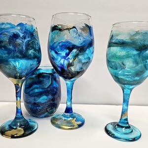 Op de afbeelding: Drie wijnglazen met blauwe en gouden abstracte ontwerpen. De glazen zijn helder en hebben een draaiende patroon van blauwe, groene en gouden verf.