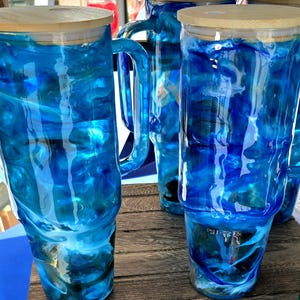Könnte beinhalten: Mehrere hohe, transparente Becher mit einem blau-weißen Marmormuster. Jeder Becher hat einen Holzdeckel, einige haben einen Griff. Die Becher werden auf einer Holzoberfläche ausgestellt.