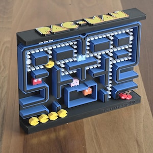 May include: A three-dimensional Pac-Man wall art piece featuring a black frame with blue maze walls, white dots, and colorful pixelated characters including Pac-Man, ghosts, and cherries. The top of the frame displays the yellow text PAC-MAN.